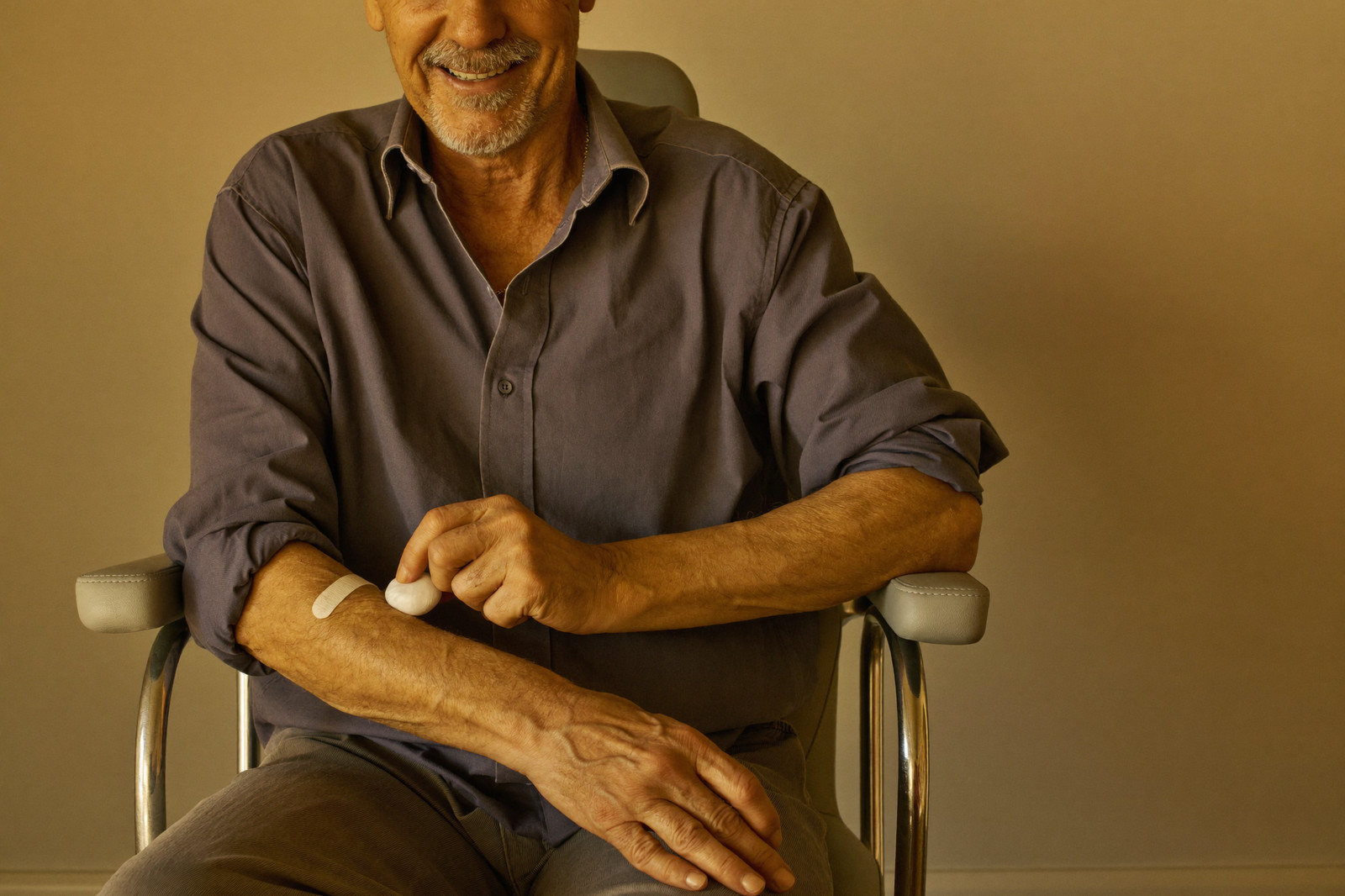 A smiling man presses a cotton ball beside a small bandage on his arm after a blood draw