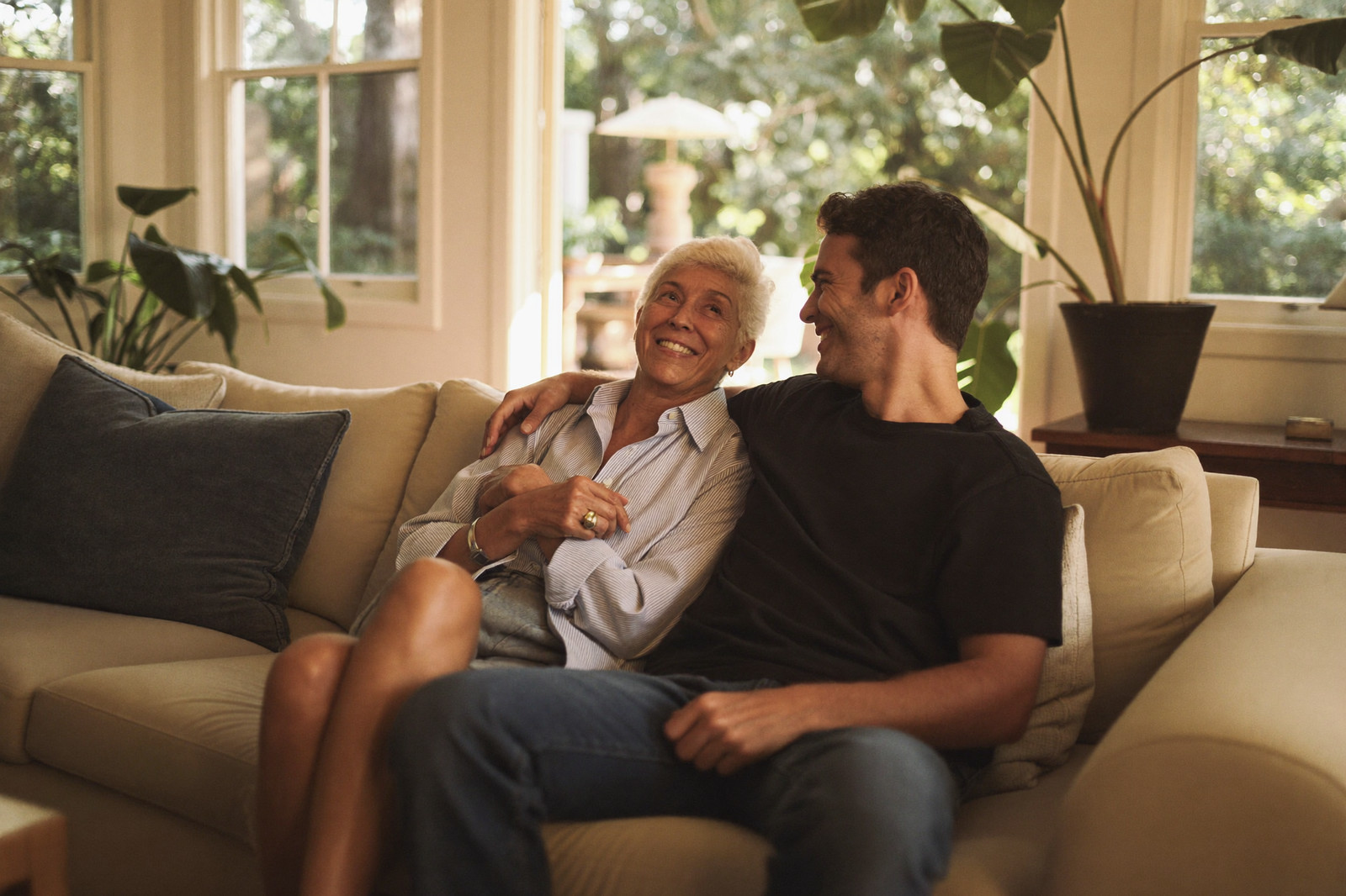 A mother and her adult son share a laugh on a sofa in a bright, plant-filled room